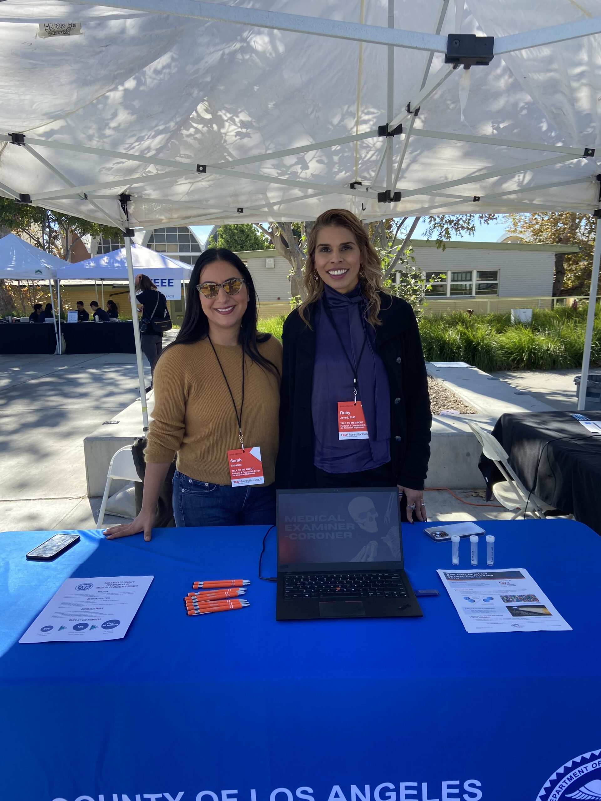 Ruby Javed with Sarah Ardelani at TEDex booth for County of Los Angeles Dept. of Medical Examiner. Tables and poster under tent.