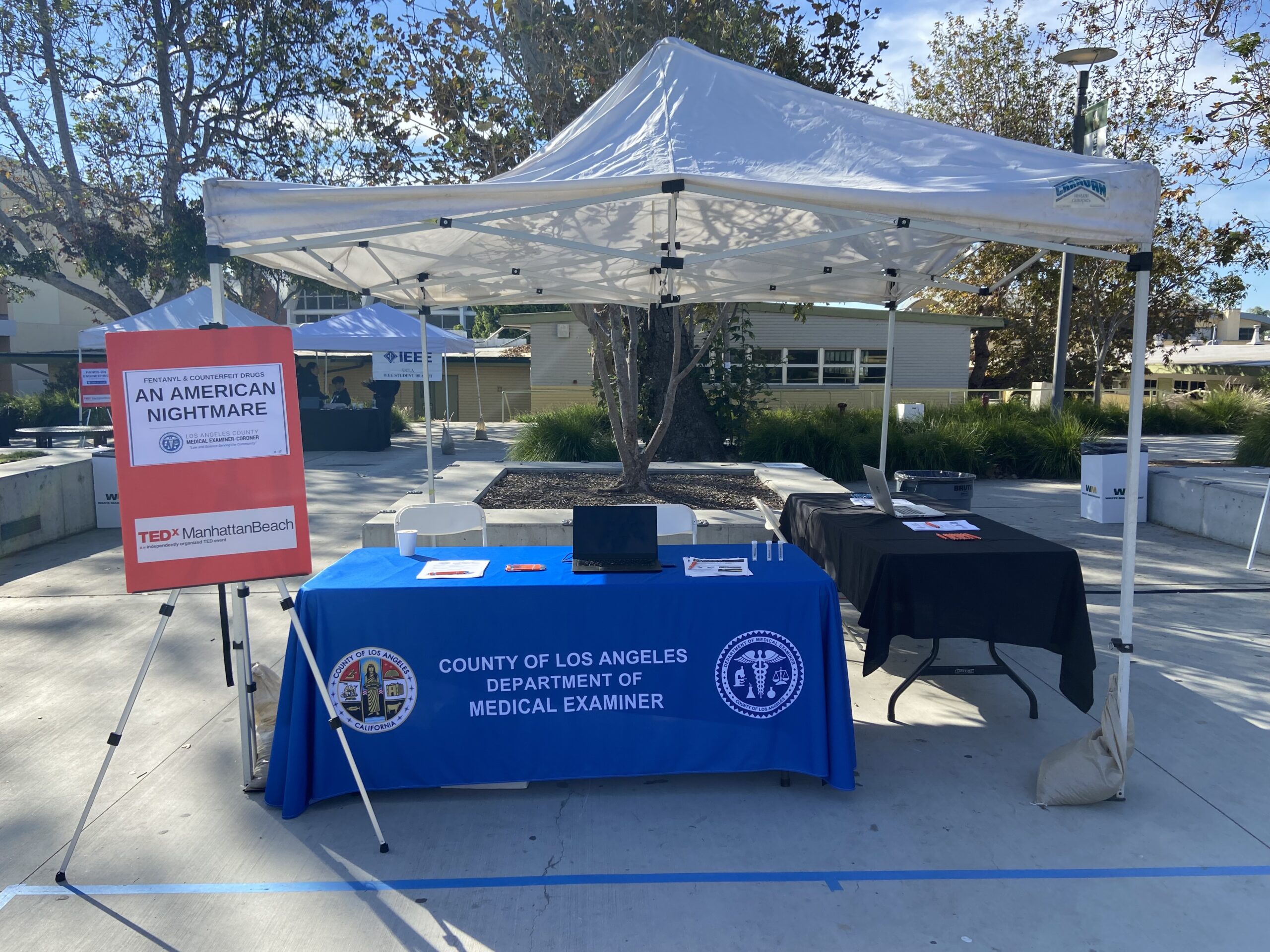 TEDex booth for County of Los Angeles Dept. of Medical Examiner. Tables and poster under tent.